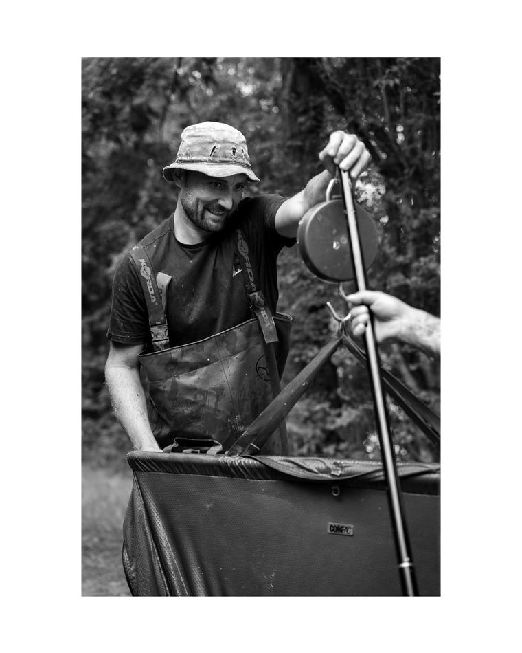 Angler weighing a carp in a sling on the bank, black and white